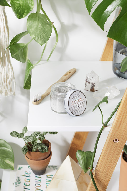 "Organic Tooth Powder and bamboo toothbrush on a wooden shelf surrounded by plants.