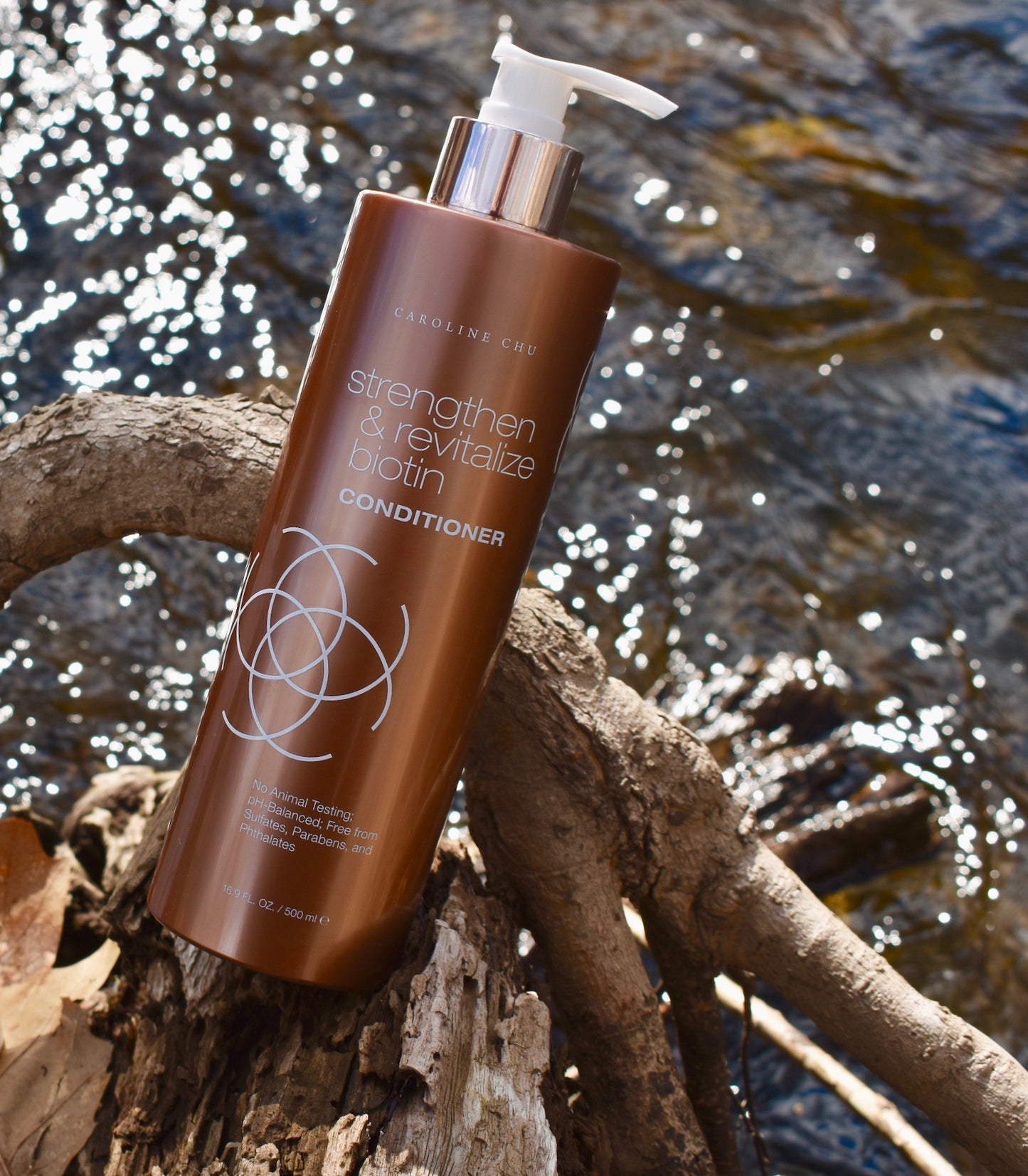 Biotin Strength & Revitalize Conditioner bottle placed outdoors near a stream, showcasing natural ingredients.